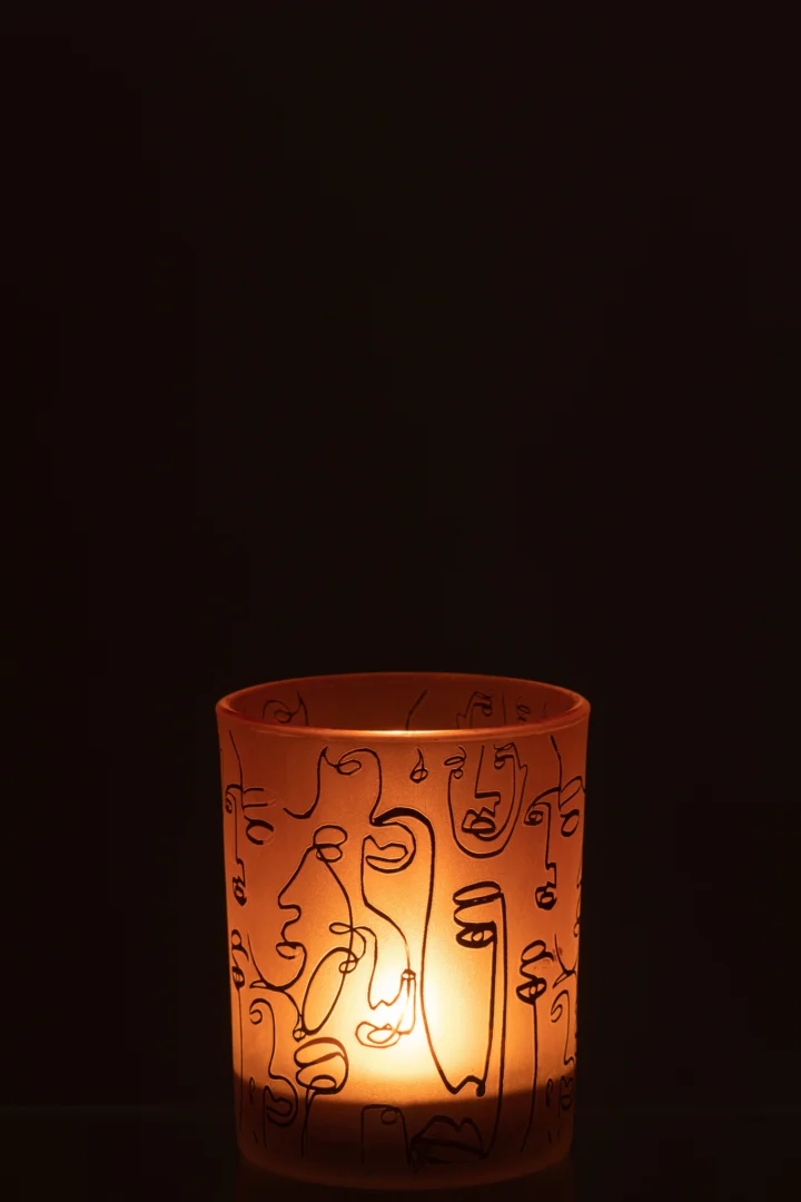 De Kaarsenhouder Orkaan Glas Terracotta Vrolijke Gezichten Medium gloeit met een brandende kaars in de oranje glazen houder, met abstracte lijnvormige gezichtsmotieven tegen een donkere achtergrond.