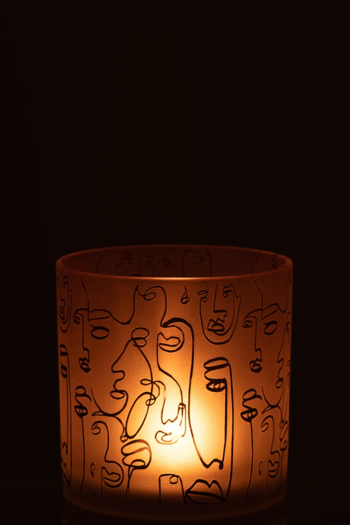 In de Kaarsenhouder Orkaan Glas Terracotta Vrolijke Gezichten Groot zit een gloeiende kaars, een ronde houder versierd met abstracte zwarte gezichtsmotieven tegen een donkere achtergrond.