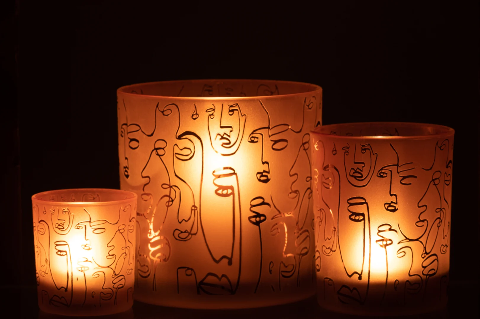 De Kaarsenhouder Orkaan Glas Terracotta Vrolijke Gezichten Groot gloeit warm op, met abstracte lijnvormige gezichtsontwerpen op matglazen houders, en creëert een uitnodigende sfeer in het donker.