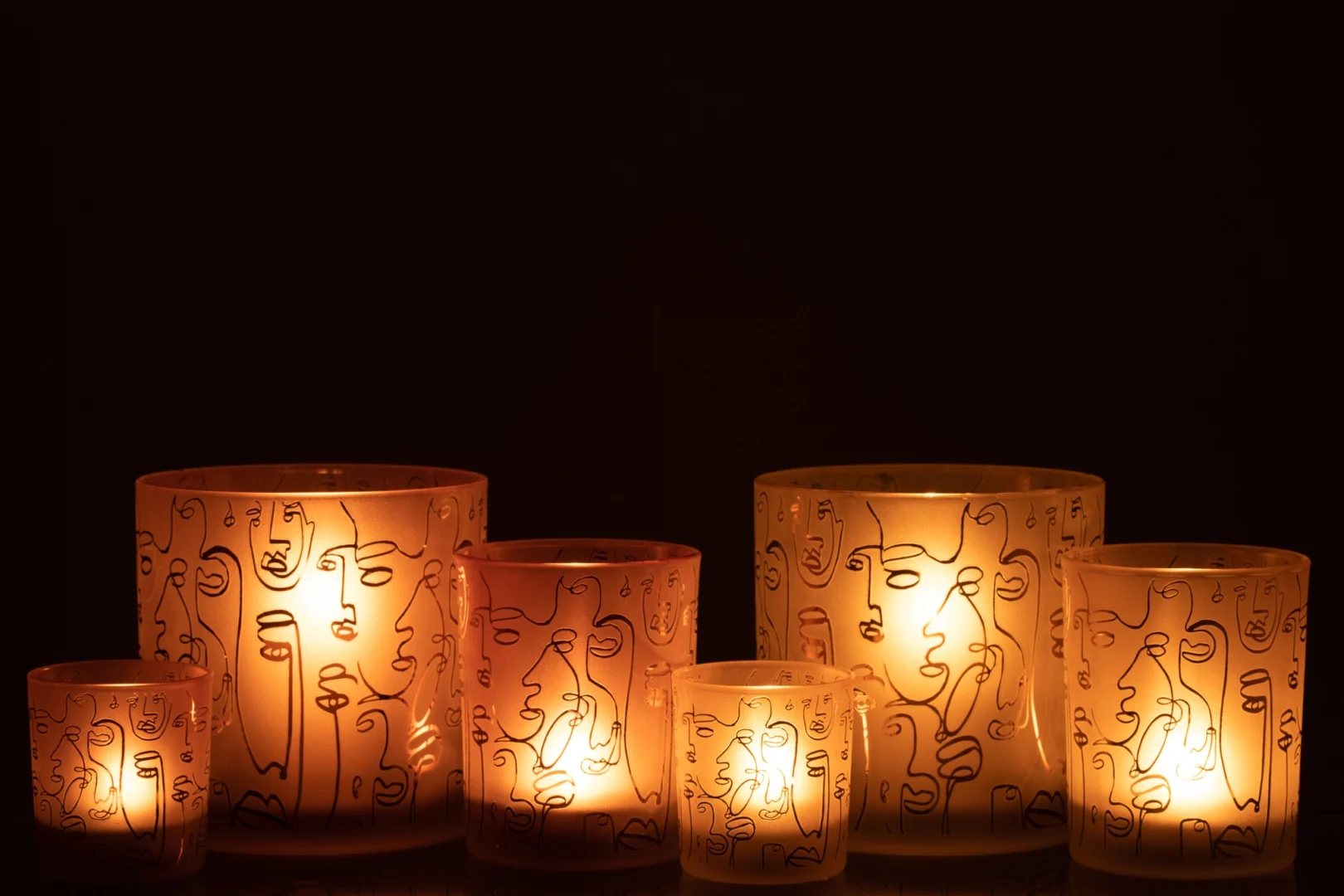 De Kaarsenhouder Orkaan Glas Terracotta Vrolijke Gezichten Groot heeft vijf gloeiende kaarsen, met abstracte gezichtsontwerpen op de houders die een gezellige en artistieke sfeer creëren tegen een donkere achtergrond.