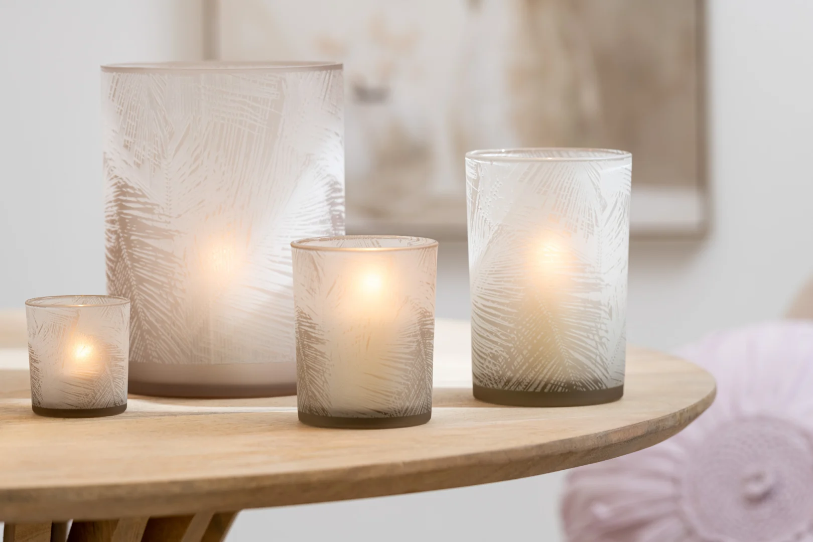 Vier Theelichthouder Bladeren Glas Beige Klein, met een subtiel bladmotief op mat glas, gloeien zacht op een ronde houten tafel en versterken de warme sfeer van een serene, neutraal getinte kamer.