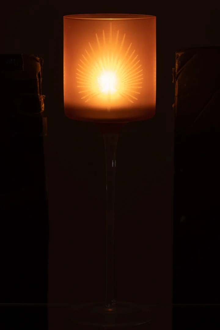 De Theelichthouder Op Voet Palmblad Glas Beige Groot werpt een warme gloed in de hoge glazen houder en creëert een stralend sterpatroon op het matglas tegen een donkere achtergrond.