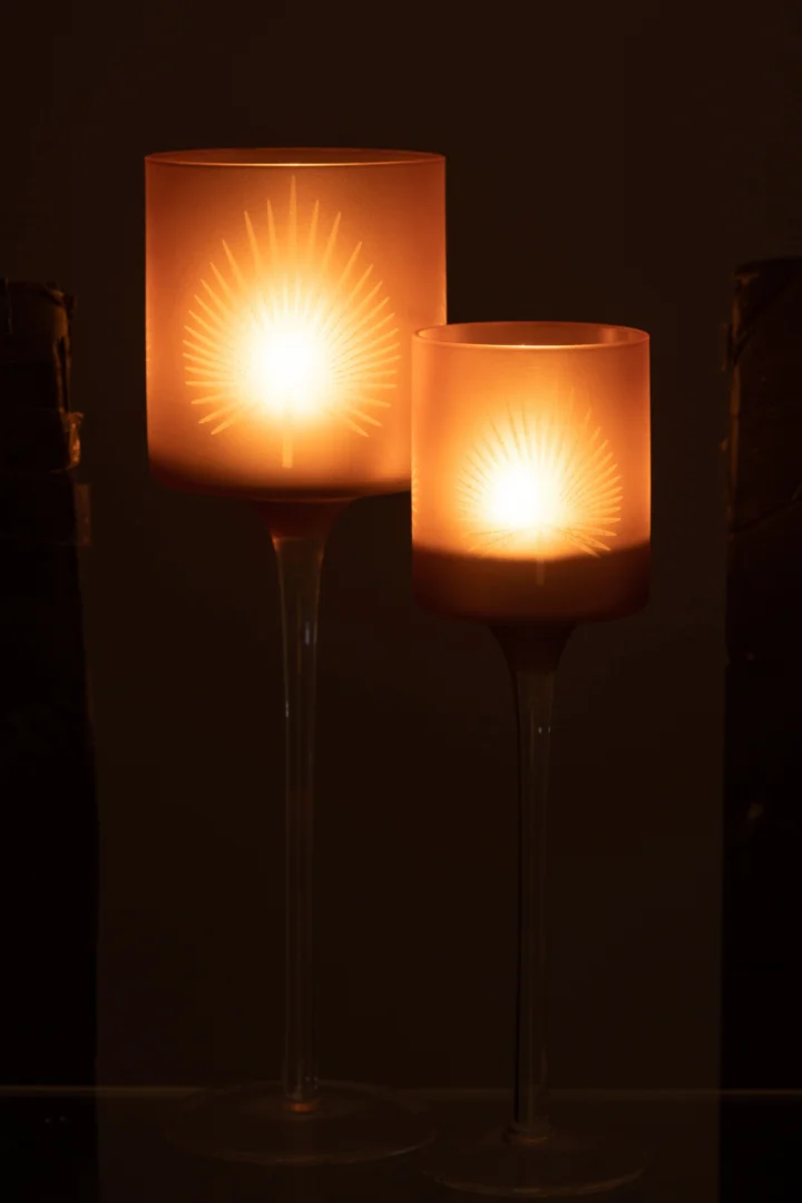 Twee Theelichthouder Op Voet Palmblad Glas Beige Groot kandelaars, elk met een gloeiende kaars erin, staan naast elkaar en werpen een sterrenpatroon over hun matglazen oppervlakken tegen een donkere achtergrond.