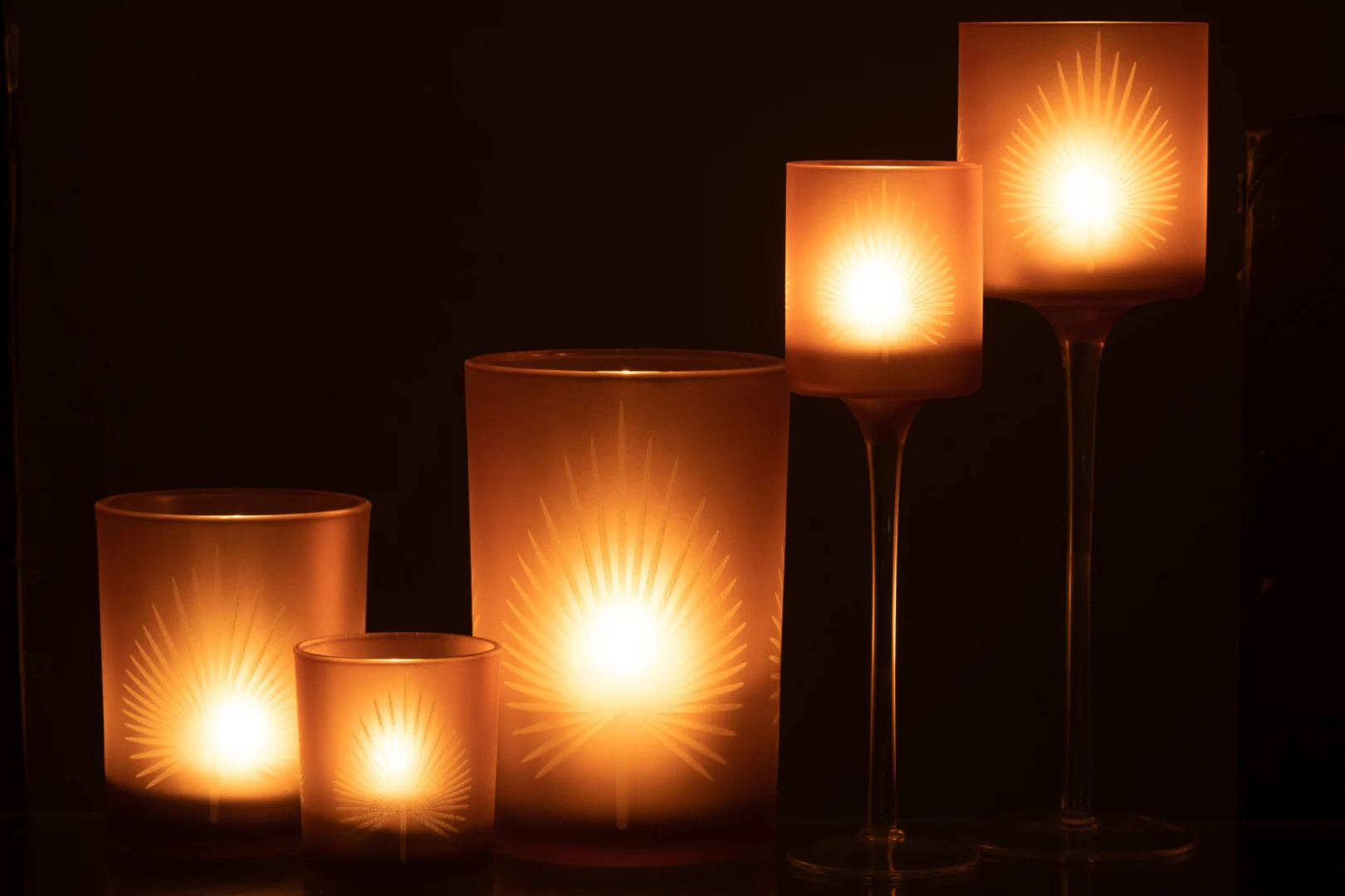 De Theelichthouder Op Voet Palmblad Glas Beige Groot straalt een warme, zachte gloed uit, terwijl de houder met sterrenpatroon een gezellige en elegante sfeer creëert in het donker.