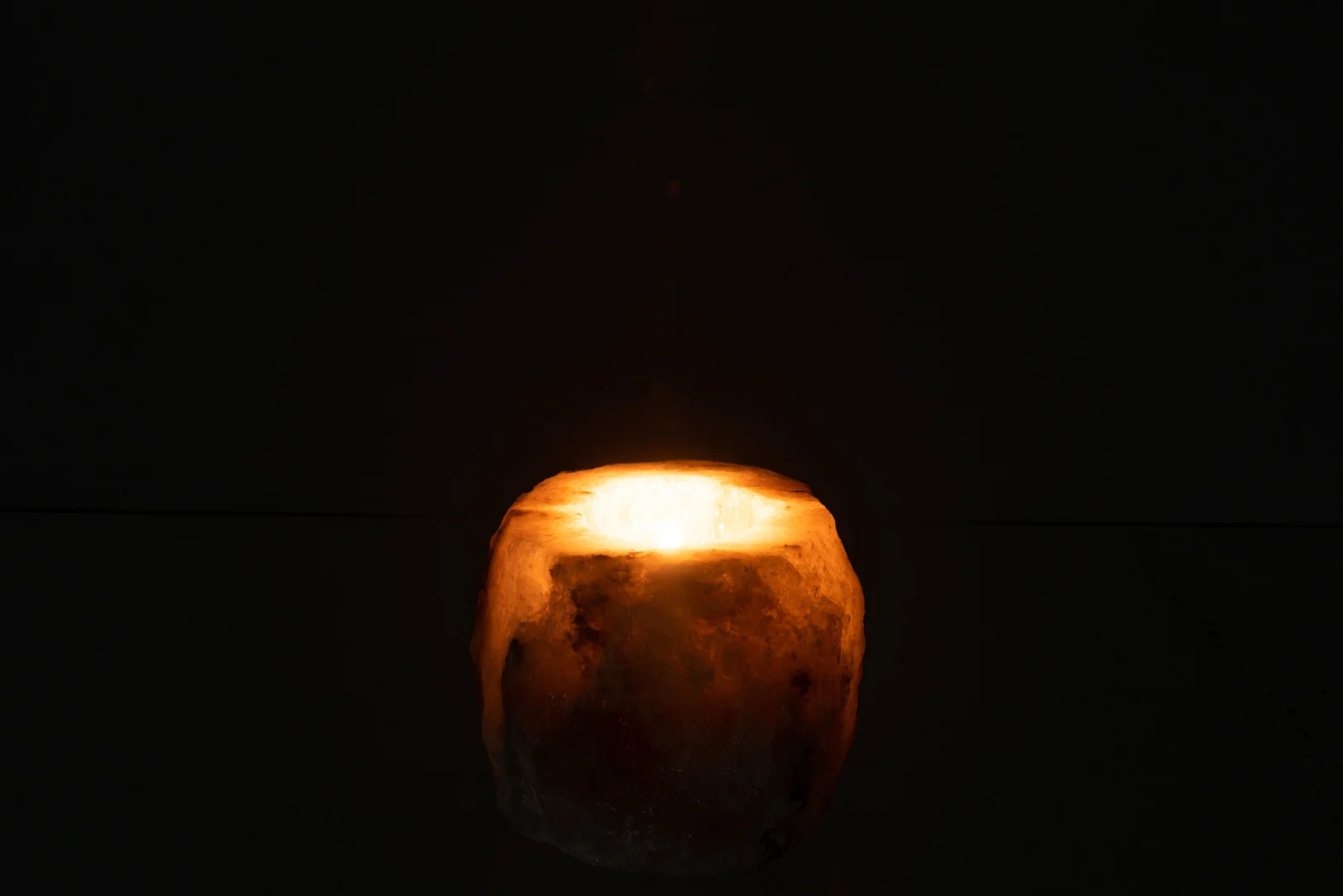 Een gloeiende kaars zit in de Kaarsenhouder Ruwe Zoutsteen Grijs, het ronde Himalayazoutontwerp met textuur werpt een zacht, warm licht in de duisternis tegen een gitzwarte achtergrond.