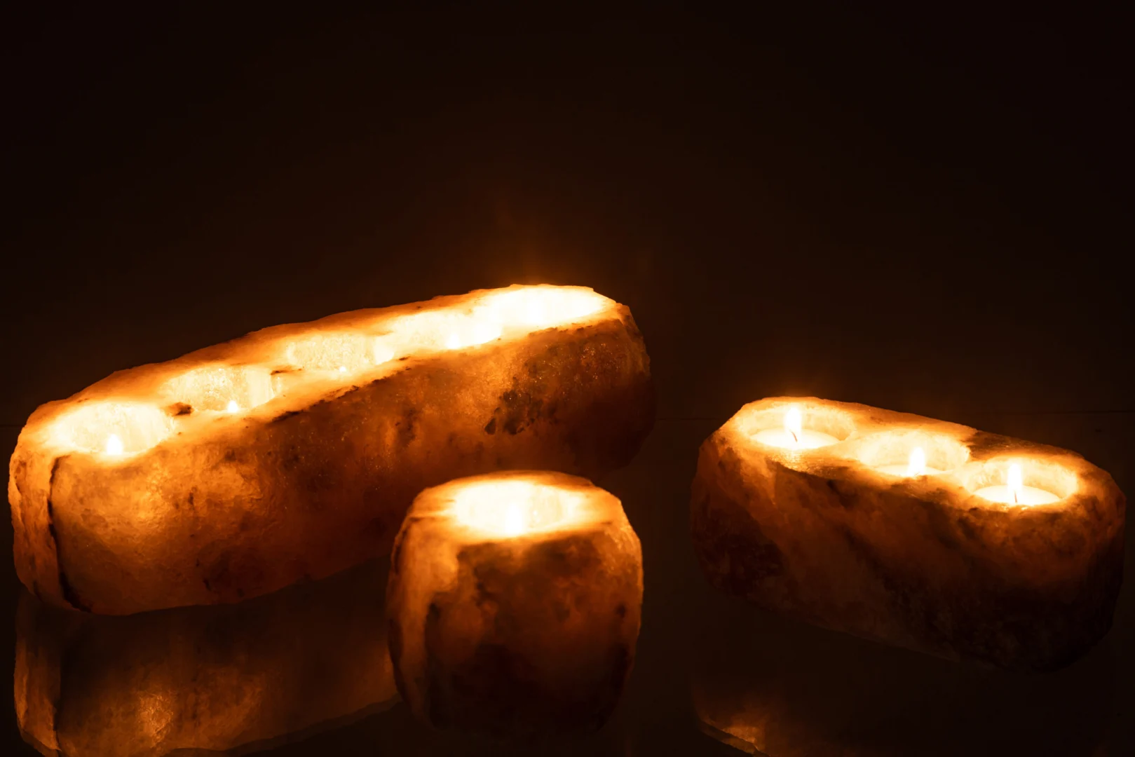 Drie Kaarsenhouder Ruwe Zoutsteen Grijs met gloeiende kaarsen staan op een reflecterend donker oppervlak, werpen een warme oranje gloed en creëren een gezellige, uitnodigende sfeer tegen de zwarte achtergrond.