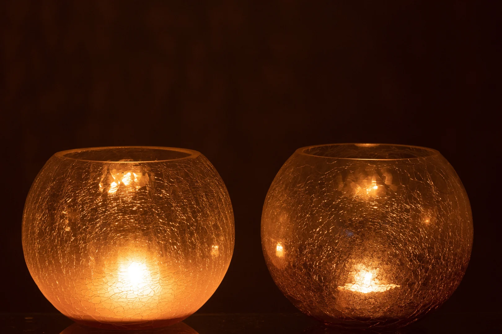 De Kaarsenhouder Orkaan Orb Craquelure Glas Paars Set 2 heeft twee paarse glazen kandelaars die een warme, zachte gloed uitstralen tegen een donkere achtergrond, waardoor een gezellige en intieme sfeer wordt gecreëerd.