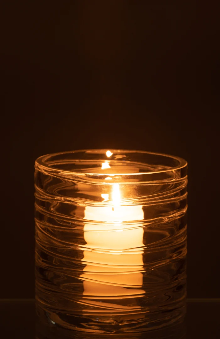 Een brandende witte kaars zit in de Kaarsenhouder Orkaan Strepen Glas Transparant Klein, de heldere glazen houder met golvende strepen werpt een warme, zachte gloed tegen een donkere achtergrond.