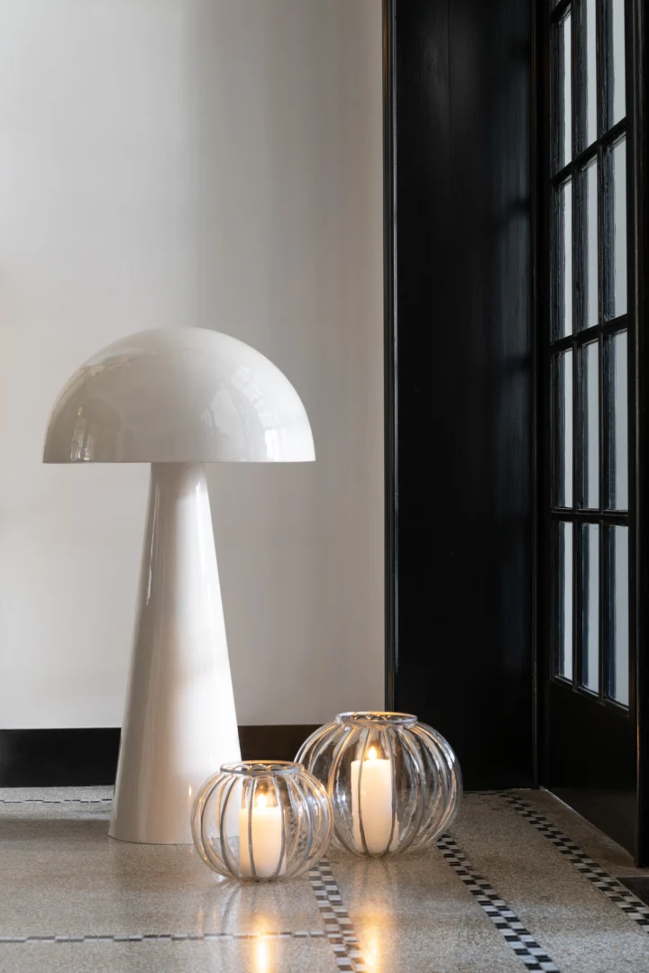 Een moderne witte paddestoelvormige lamp staat naast twee verlichte Kaarsenhouder Orkaan Strepen Glas Transparant Klein kandelaars op een betegelde vloer bij een zwart omlijst raam.