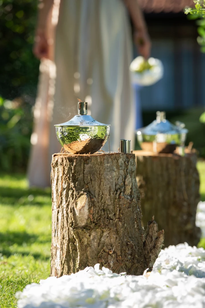 De Olielamp Roma RVS Zilver rust buiten op een boomstronk, omringd door verspreide witte bloemblaadjes. Op de achtergrond draagt een vage figuur die bloemen vasthoudt bij aan de sfeer in de tuin.