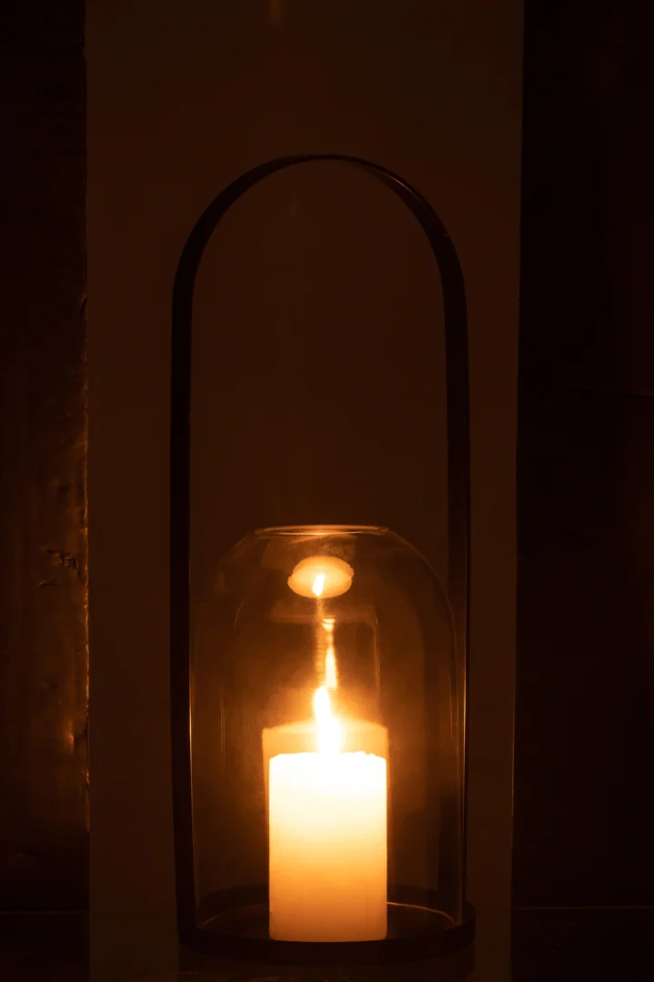 De Kaarsenhouder Orkaan Half Rond Metaal Glas Zwart Groot houdt een gloeiende witte kaars vast en werpt een warm, zacht licht door de glazen koepel en het zwarte metalen frame in een verduisterde kamer.