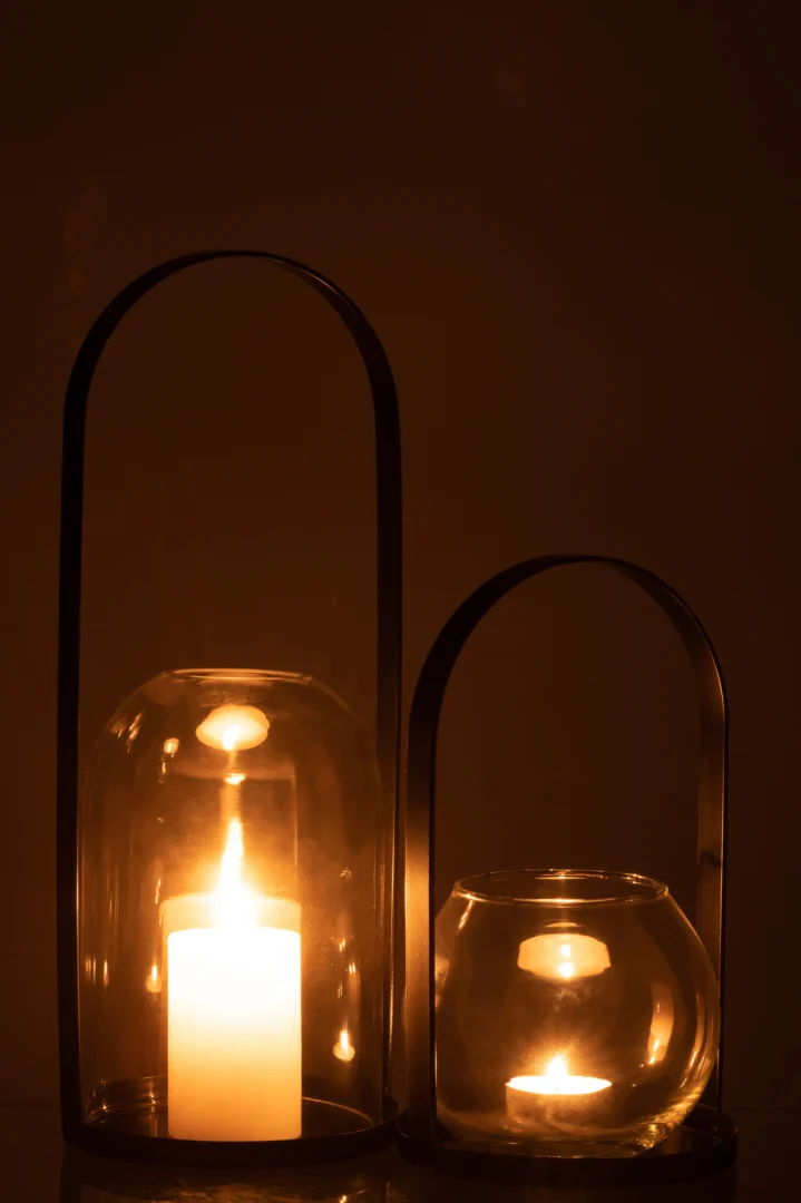 Een hoge kaars gloeit in de Kaarsenhouder Orkaan Half Rond Metaal Glas Zwart Groot, zijn glazen en metalen koepel werpt warm licht in een donkere kamer naast een kortere kaars in een ronde glazen houder.