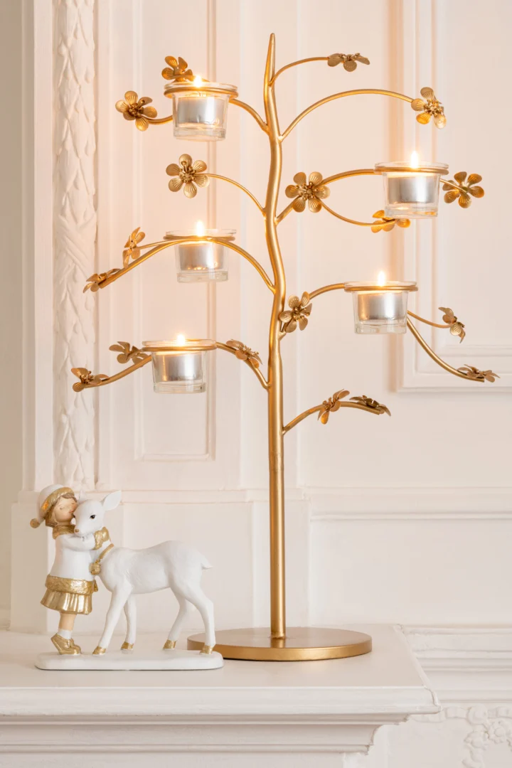 De Kaarsenhouder Tafel Metaal Goud, een goudmetalen kandelaar in de vorm van een boom met brandende kaarsen, staat op een schoorsteenmantel naast een wit en goudkleurig beeldje van een meisje met een hert tegen een sierlijke witte muur.