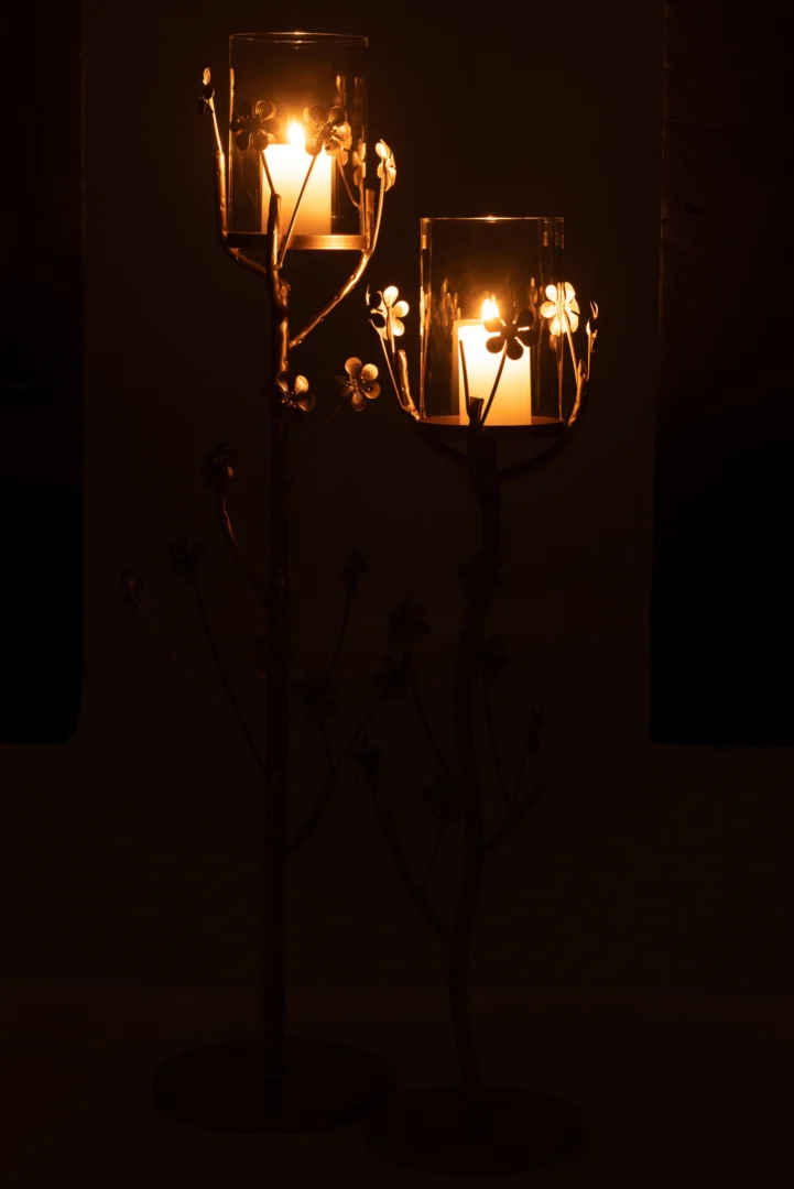 Twee brandende kaarsen in de Kaarsenhouder Vloer Metaal Goud Groot werpen een warme gloed in een donkere kamer, hun glazen houders gemonteerd op decoratieve goudmetalen standaards met elegante bloemmotieven.