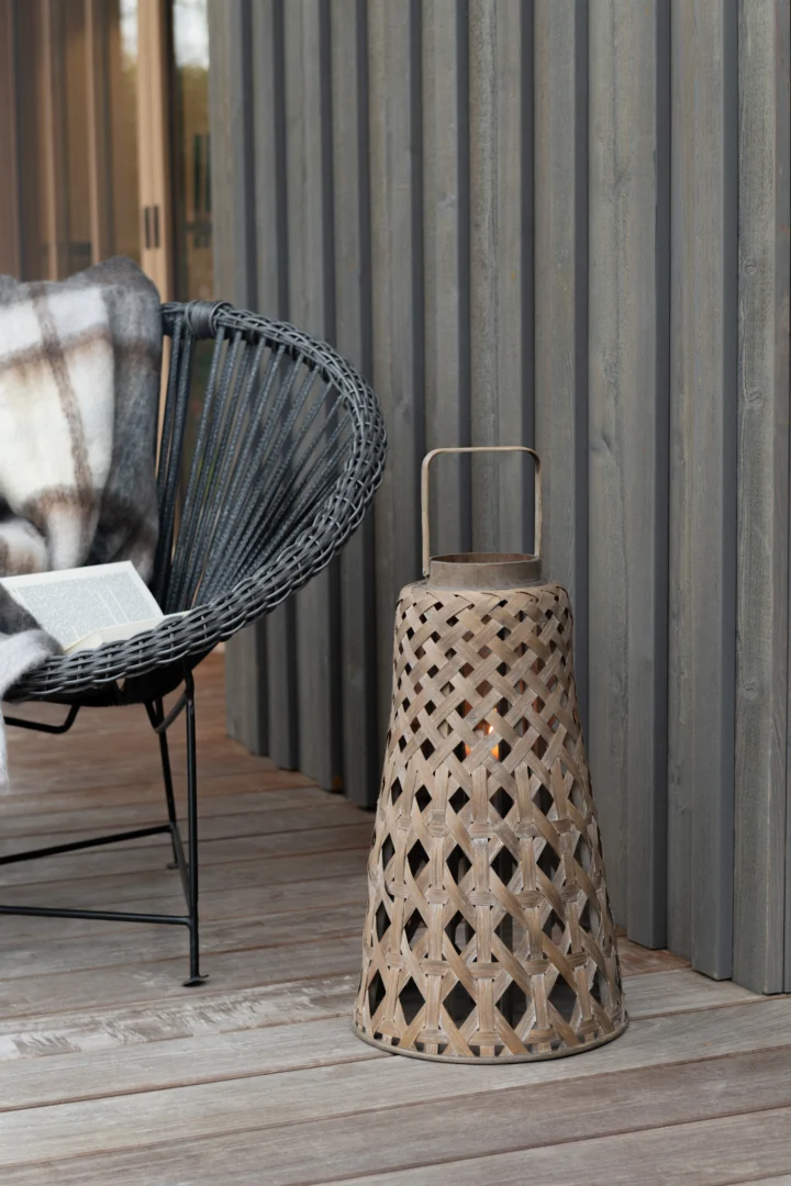 Een geweven stoel met een geruite deken en een open boek staat naast de Lantaarn Bamboe Natuurlijk op een houten dek, naast een muur met verticale houten lambrisering.
