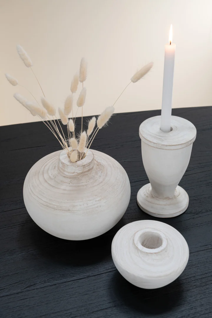 Een grote witte houten kandelaar (Kaarsenhouder Hout Wit Groot) met een brandende witte kaars staat op een donkere tafel naast een ronde vaas met gedroogd gras en een kleine ronde schaal met deksel ernaast.