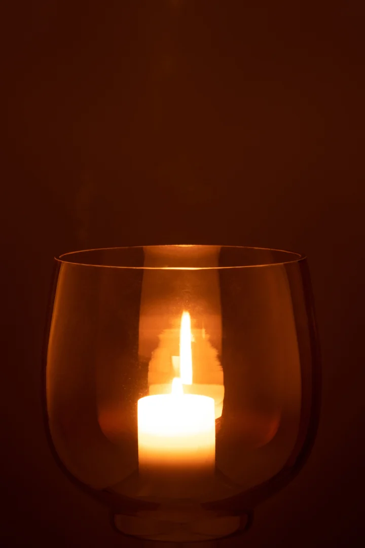 Een brandende kaars gloeit warm in de Kaarsenhouder Orkaan Glas Rook Geel Zilver Groot, de ronde glazen houder werpt zachte reflecties in het schemerige licht.