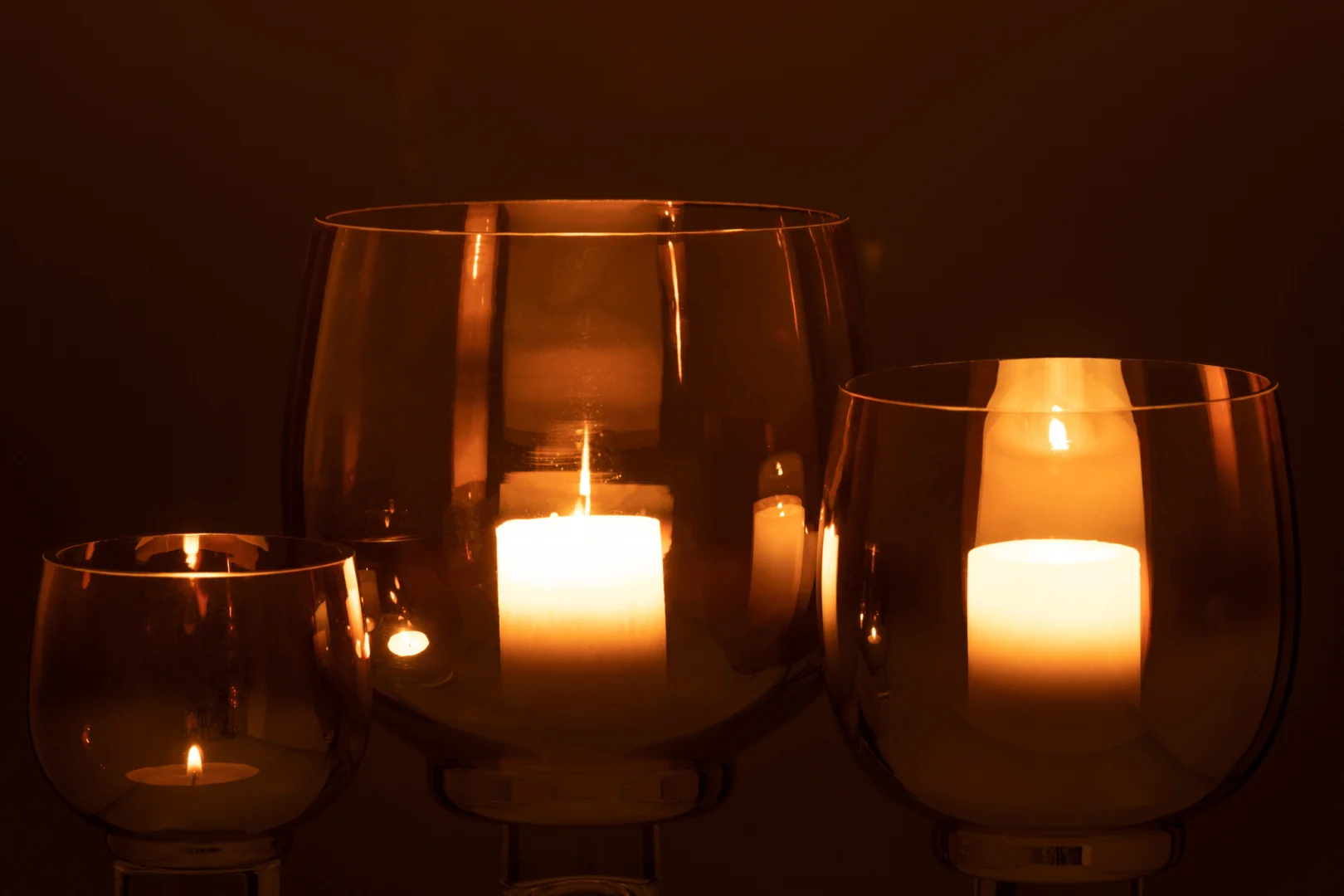 Een Kaarsenhouder Orkaan Glas Rook Geel Zilver Groot houdt een brandende witte kaars vast en werpt een warme, zachte gloed en zachte reflecties op het glas tegen een donkerbruine achtergrond.