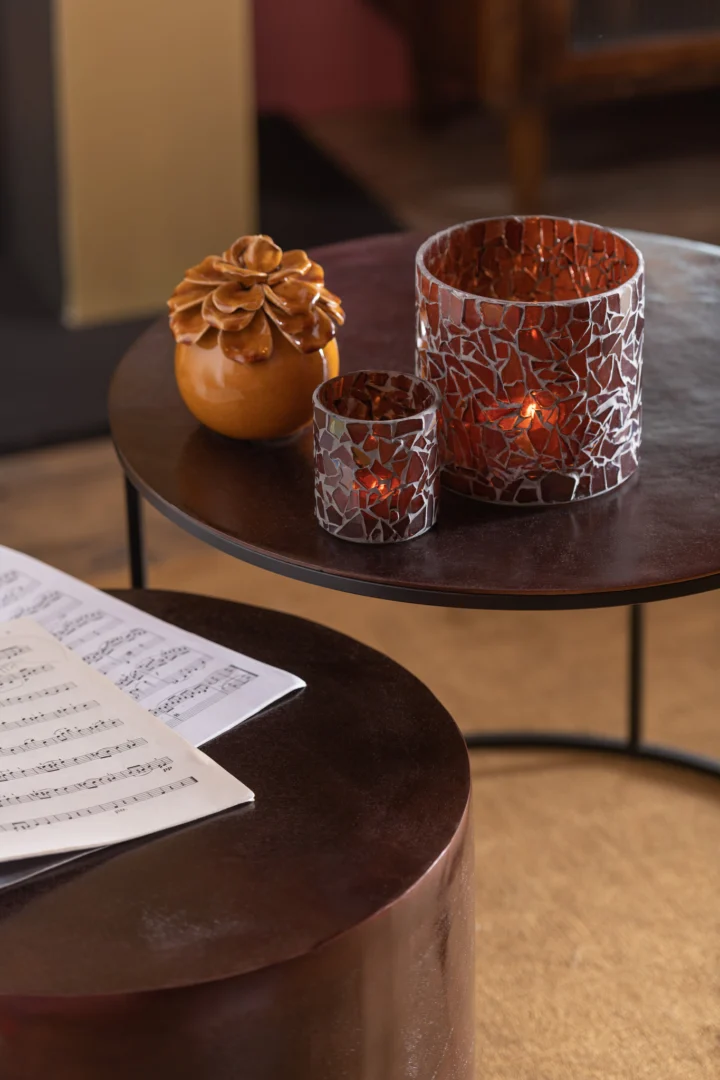 Twee ronde tafels met een bruine afwerking. Op de bovenste tafel staan twee Theelichthouder Mozaïekglas Bruin Klein kandelaars met brandende kaarsen en een bloem van keramiek. Op de onderste tafel ligt bladmuziek. Warme, gezellige binnenverlichting.