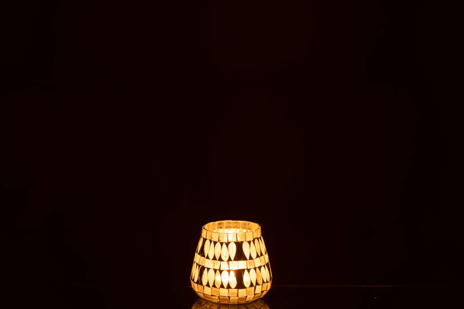 De Theelicht Mozaïek Glas Beige Klein straalt een warme, zachte gloed uit met zijn kleine mozaïekglasontwerp, dat prachtig afsteekt tegen een donkere zwarte achtergrond.