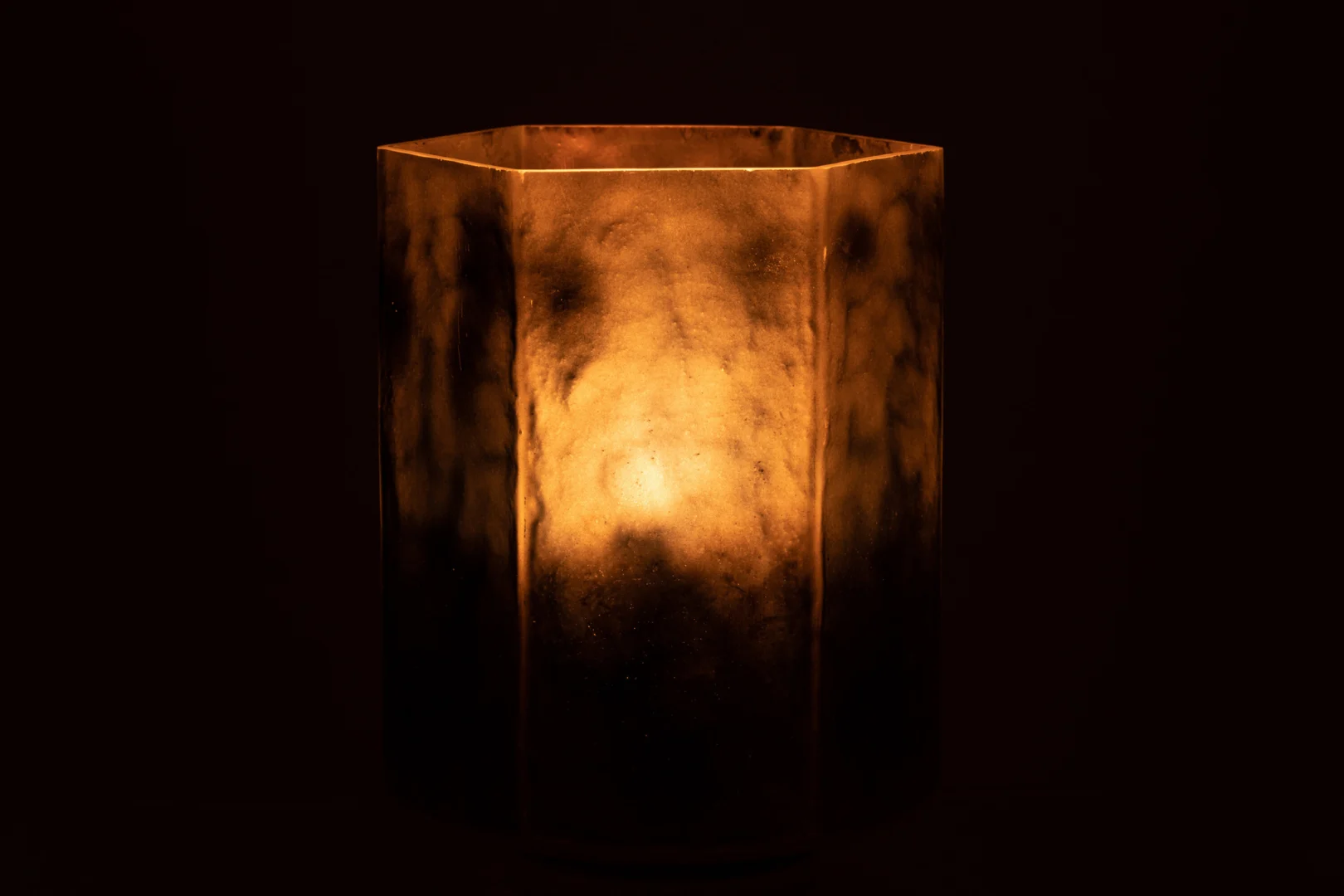 De Kaarsenhouder Orkaan Hexagoon Glas Grijs Groot is een grote, zeshoekige glazen lantaarn met een gemarmerd grijs, halfdoorschijnend oppervlak dat in het donker een warme, gouden gloed afgeeft.