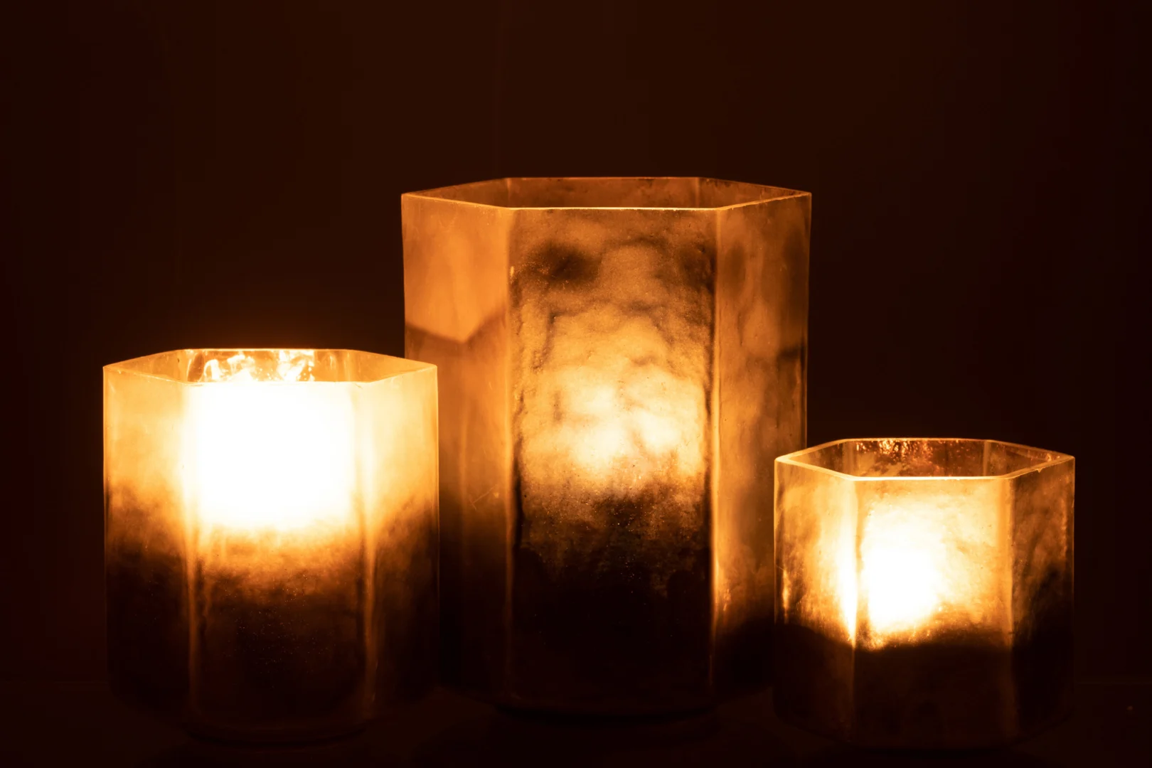 De Kaarsenhouder Orkaan Hexagoon Glas Grijs Groot is een grote zeshoekige glazen kandelaar die warm gloeit en sfeervol licht werpt tegen een donkere achtergrond.