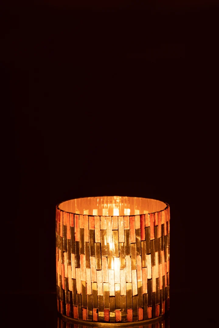 De Orkaan Chic Glas Bruin/Goud Medium kaars gloeit warm in de cilindrische houder met een goudkleurig, metallic, baksteenachtig patroon tegen een donkere achtergrond.