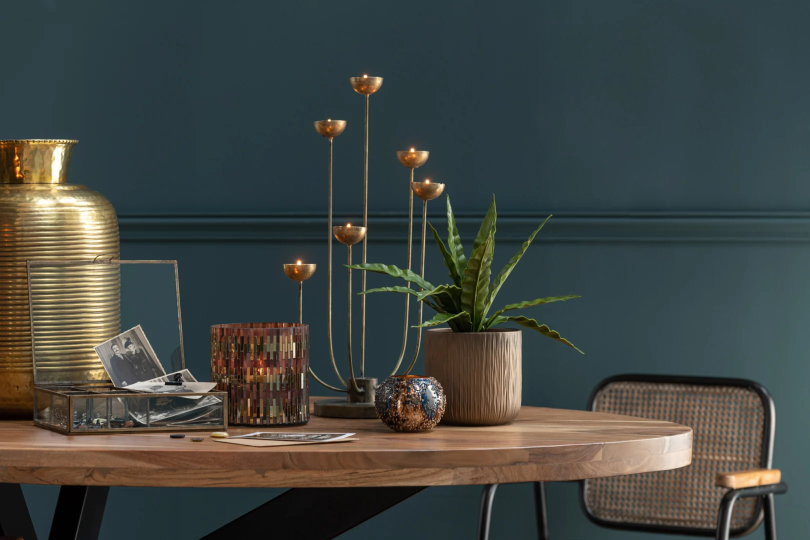 Op een ronde houten tafel staan de Orkaan Chic Glas Bruin/Goud Medium vaas, een moderne gouden kandelaar, een potplant, kandelaars met kleurrijke patronen, vintage zwart-wit foto's en een stoel met netrug tegen een donkerblauwe muur.