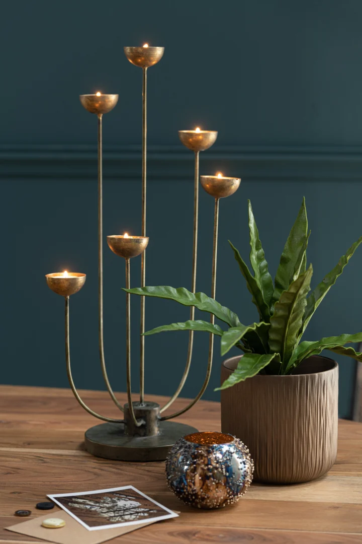 Een gouden metalen kandelaar met vijf brandende kaarsen staat op een houten tafel naast een groene potplant, foto's en Theelichthouder Lise Glas Bruin Goud Klein, allemaal tegen een donkergroengekleurde muur.