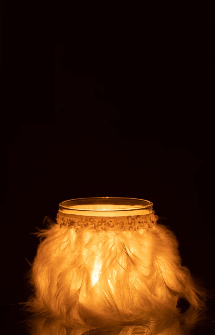 Theelichthouder Veren Wit Glas Transparant Middel: Een gloeiende kaars in een transparante glazen houder met witte veren en pailletten, tegen een donkere achtergrond. Het warme licht verlicht de veren zachtjes en creëert een gezellige sfeer.