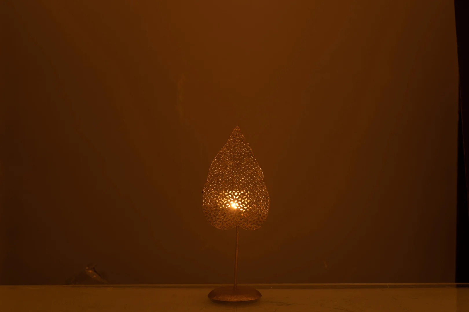 De Theelichthouder Blad Metaal Goud Klein, een kleine goudmetalen waxinelichthouder met een geperforeerd bladontwerp, zit op een oppervlak en werpt warm licht en ingewikkelde schaduwen tegen een effen bruine achtergrond.