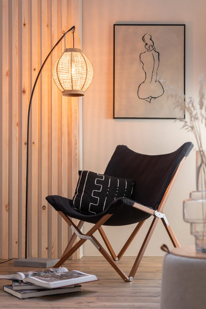 Een gezellig leeshoekje met een moderne zwarte stoel, een kussen met patroon, een open boek op de grond en warme verlichting van de Lantaarn Bamboe Natuurlijk vloerlamp. Een minimalistisch lijnkunstportret maakt de ontspannen sfeer compleet.