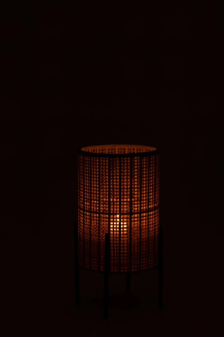 De Kaarsenhouder Lantaarn Op Voet Bamboe Groot is een geweven, cilindervormige lantaarn die een warme, sfeervolle gloed uitstraalt door het gestructureerde bamboepatroon, dat afsteekt tegen een donker oppervlak en een zwarte achtergrond.