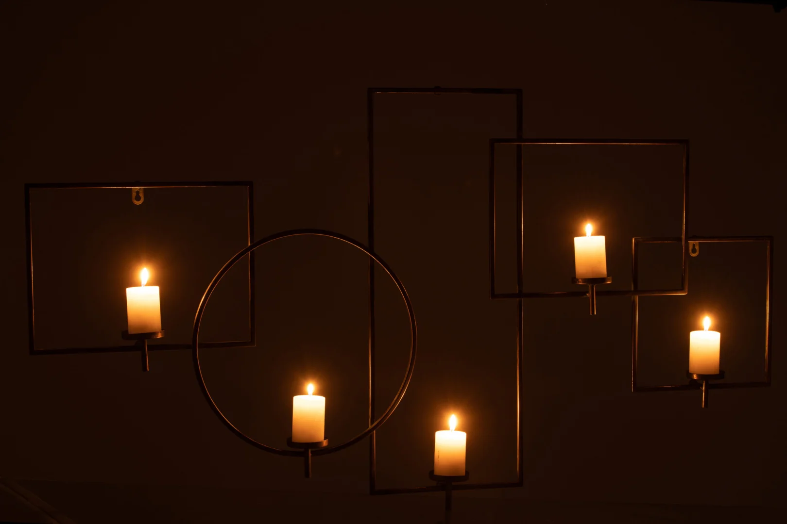 De Wanddecoratie Kaarsenhouder Metaal Goud heeft vijf brandende kaarsen in minimalistische gouden metalen frames - rechthoeken en een cirkel - tegen een donkere achtergrond voor een warme, sfeervolle gloed.