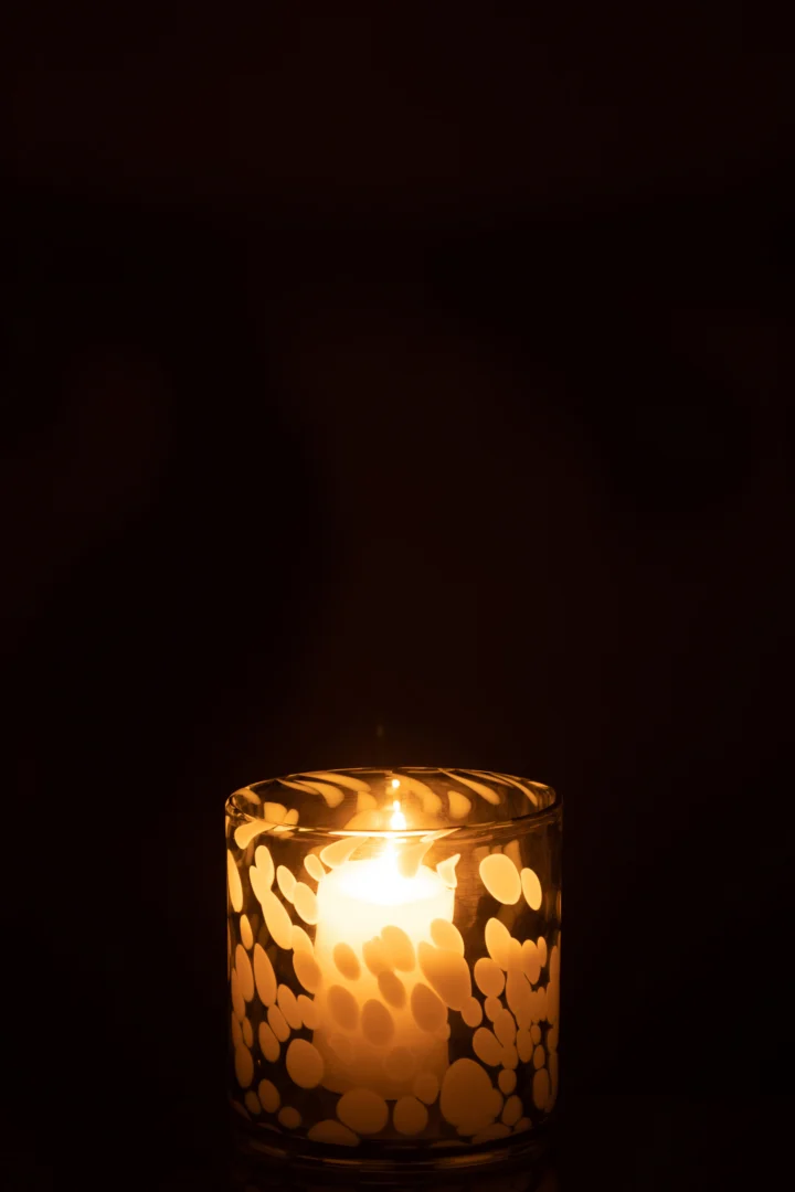 De Kaarsenhouder Orkaan Dot Glas Grijs Wit Klein verlicht het donker zachtjes met zijn warme gloed, met een brandende kaars in een glazen houder versierd met witte, cirkelvormige patronen.