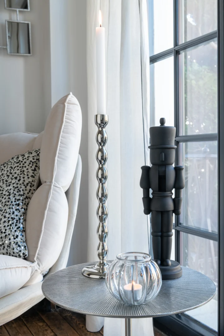 Op een ronde tafel naast een witte bank staat de Kandelaar Bol Aluminium Zilver Groot met een brandende witte kaars, een zwart notenkrakerfiguurtje en een glazen waxinelichthouder met kaars, bij een groot raam met witte gordijnen.