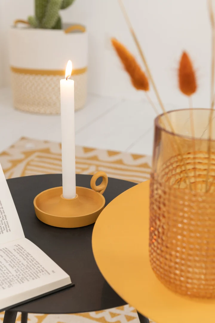 Een brandende witte kaars in de Kaarsenhouder Schaal Rond Aluminium Oker staat op een zwarte tafel bij een open boek. Een amberkleurig glas met textuur, gedroogde grassen op een geel dienblad en een plant op de achtergrond maken het tafereel compleet.