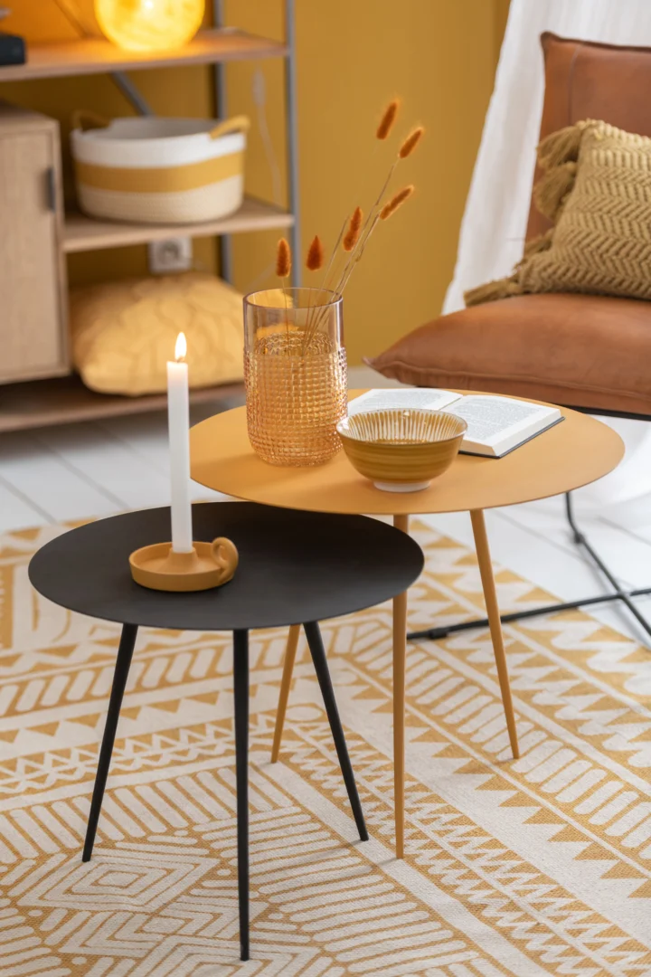 Een gezellige kamer met een gele inrichting heeft twee nesttafels, een zwarte en een mosterdgele, gestyled met de Kaarsenhouder Schaal Rond Aluminium Oker kandelaar, een vaas met gedroogde bloemen, een schaal en een open boek op een vloerkleed met patroon.