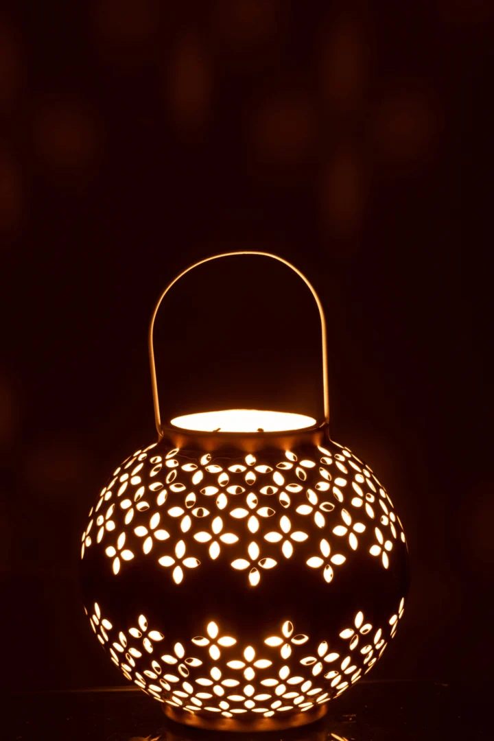 De Kaarsenhouder Lantaarnbal Geperforeerd IJzer Oker is een ronde metalen lantaarn met bloemvormige uitsnijdingen, een gebogen handvat en straalt een warme, sfeervolle gloed uit die ingewikkelde patronen in het donker werpt.