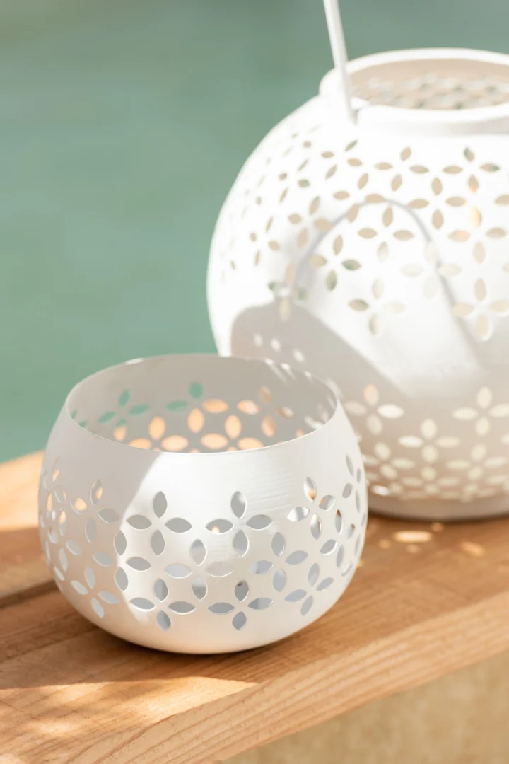 Twee witte Theelichthouder Geperforeerd IJzer kandelaars met uitgesneden bloemmotieven rusten op een houten oppervlak. Zacht zonlicht werpt decoratieve schaduwen tegen een vage lichtblauwe achtergrond.