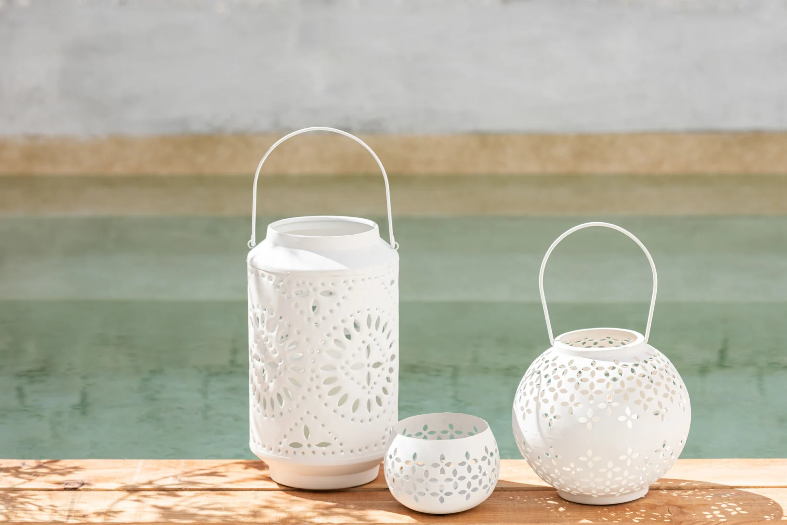 Drie witte Theelichthouder Geperforeerd IJzer lantaarns staan op een houten oppervlak aan het water. Hun ingewikkelde uitgesneden ontwerpen werpen zachte schaduwen in het zonlicht.