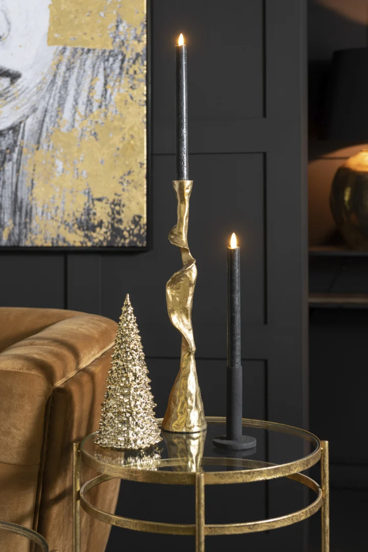 Op een kleine ronde gouden tafel staat de Kaarsenhouder Ozy Aluminium Goud Set 3 met een zwarte taps toelopende kaars en bijpassend decor, tegen donkere muren met goud geaccentueerde details.