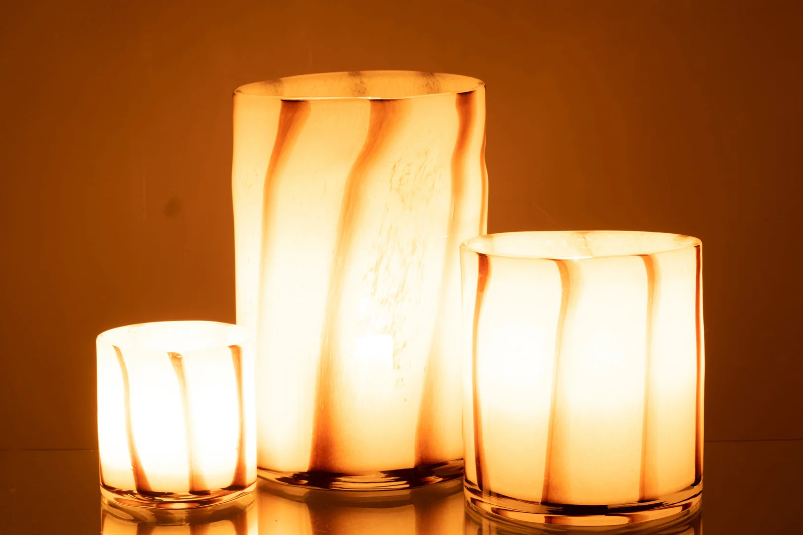 De Kaarsenhouder Orkaan Strepen Safari Glas Wit Bruin Groot heeft golvende bruine en witte strepen die samen een gezellige, uitnodigende sfeer creëren tegen een warme amberkleurige achtergrond.