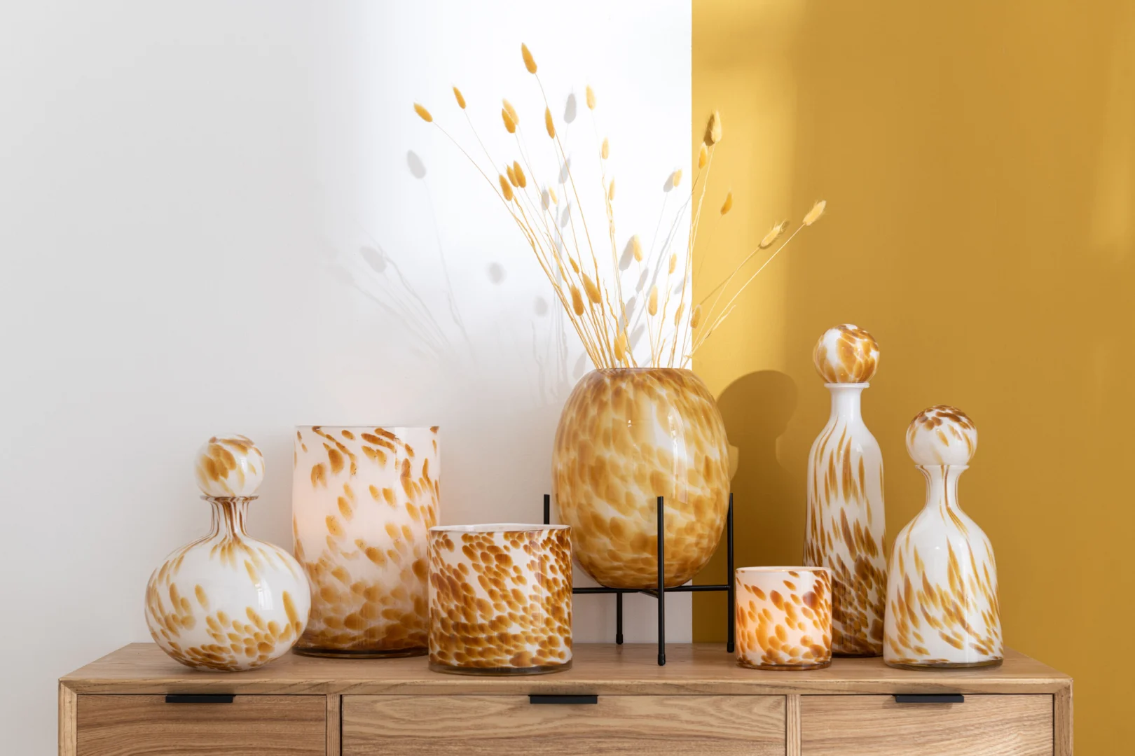 De Kaarsenhouder Orkaan Dot Glas Wit Oker Medium, met wit en oker gespikkeld glas, staat op een houten kast tegen witte en mosterdgele muren, met gedroogde grassen in de hoge vorm.