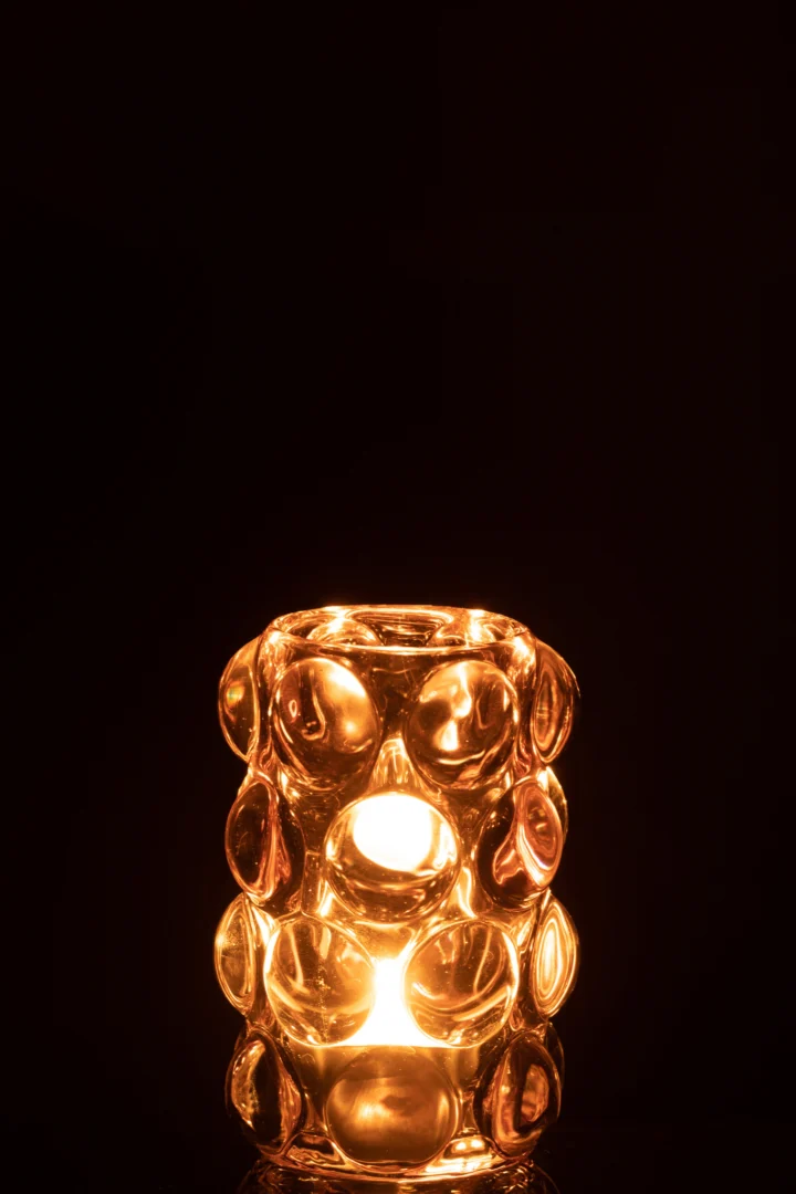 De Theelichthouder Bollie Glas Roze heeft een gloeiende kaars in een roze glazen houder met verhoogde cirkelvormige patronen, die een warm, gouden licht werpt tegen een donkere achtergrond.