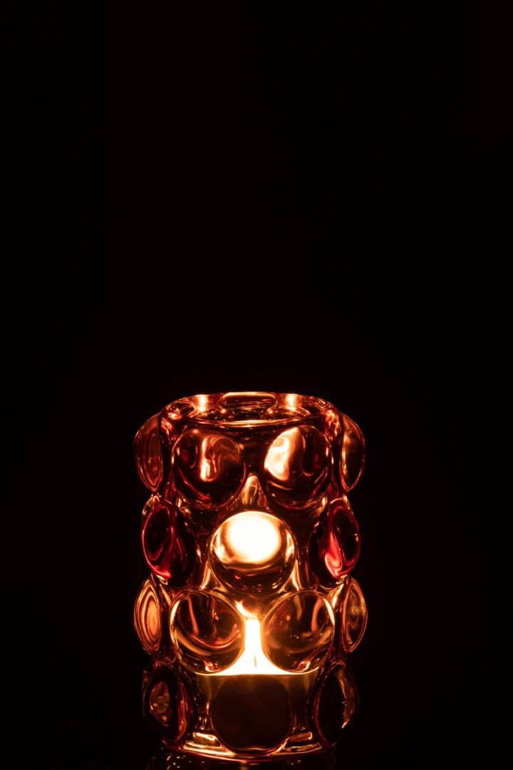 De Theelichthouder Bollie Glas Rood heeft een enkele kaars die gloeit in een rood glazen houder met cirkelvormige patronen, die warm, gouden licht werpt tegen een donkere achtergrond.