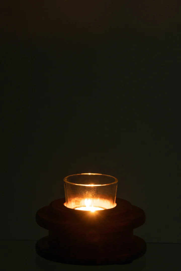 De Kaarsenhouder Spoel Hout Bruin heeft een kleine kaars in een heldere glazen houder op een donkere houten voet, die licht om zich heen werpt tegen een donkere achtergrond.