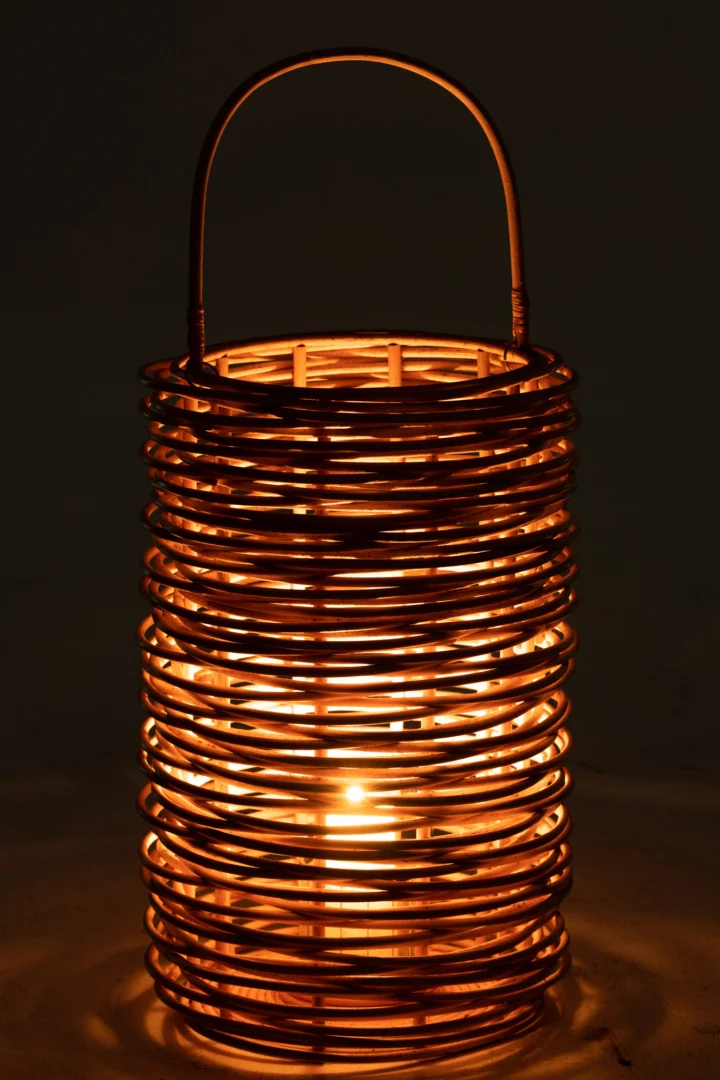 De Lantaarn Isabelle Rotan Natuurlijk is een cilindervormige lantaarn met een handvat, gemaakt van natuurlijk rotan en van binnenuit verlicht door een warm, gloeiend licht.