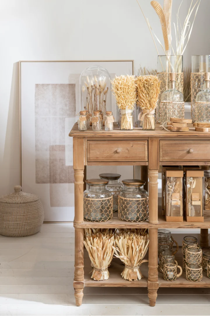 Een houten tafel met potten en mokken staat in een lichte, neutrale kamer, gedecoreerd met droogbloemen, ingelijste kunst, een geweven poef en de Kaarsenhouder Orkaan Glas Staaf Brei Transparant Medium voor een natuurlijke, rustieke touch.