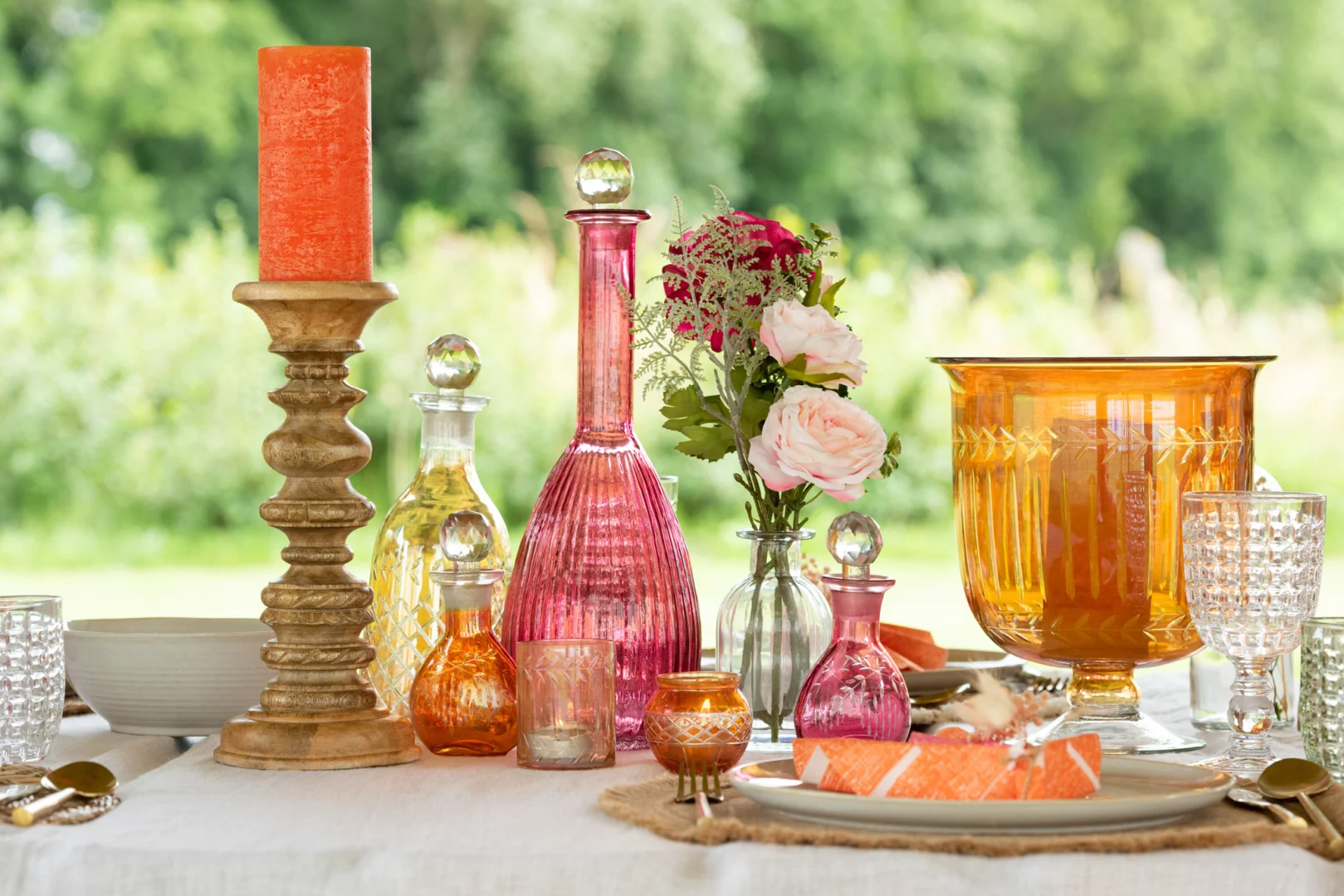 Een levendige tafelschikking met kleurrijke glazen flessen, een roze vaas met bloemen, amberkleurig glaswerk, borden en Theelichthouder Bladeren Glas Rond Mix Set van 3 kandelaars op een wit tafelkleed.